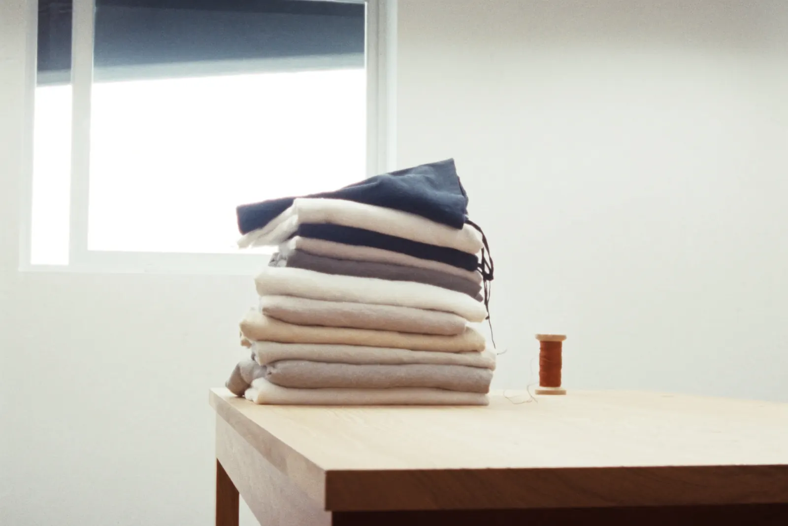 A stack of folded cloth in cream, grey and navy on an oak table, with a spool of orange thread beside it