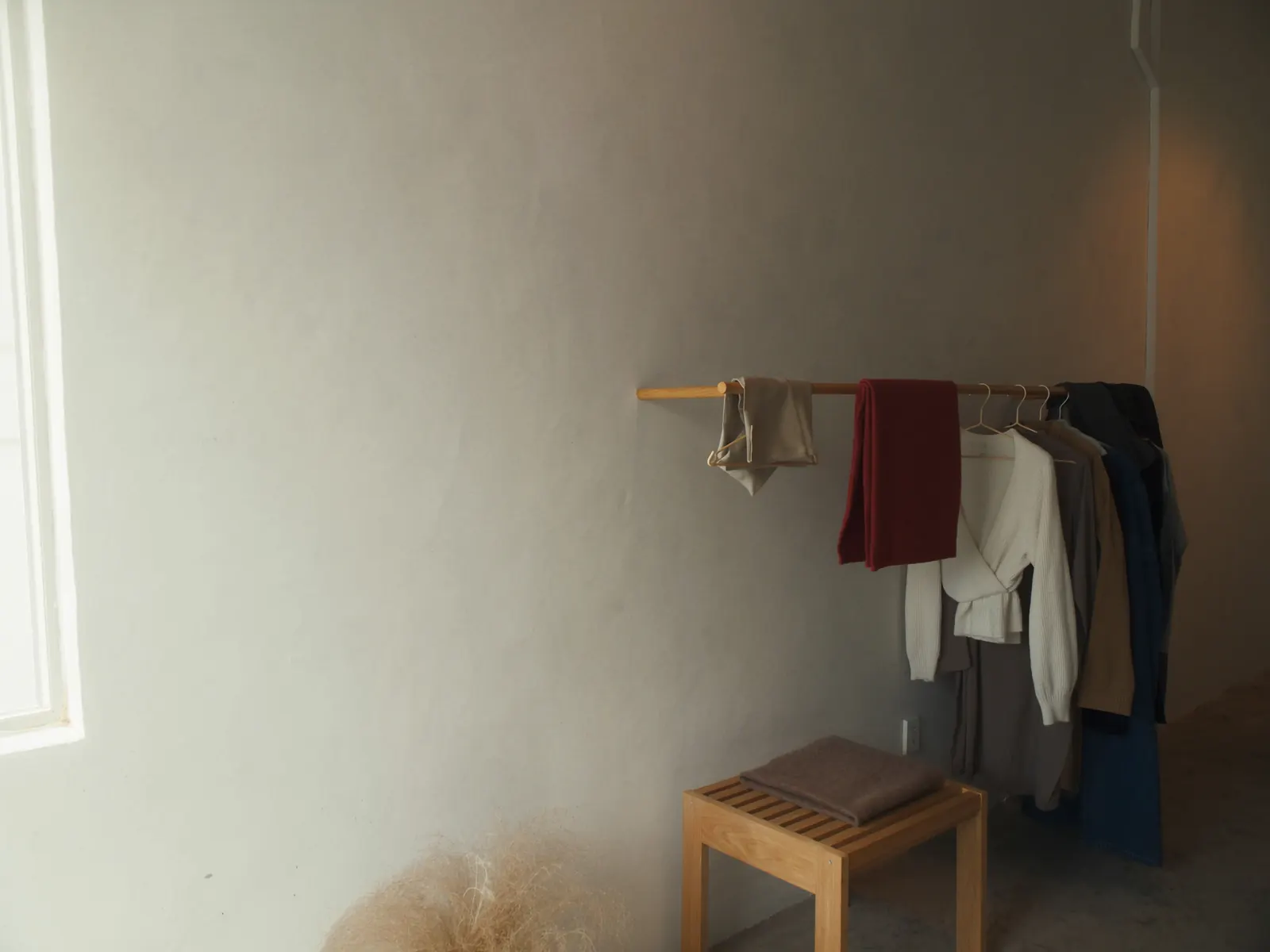 A wooden clothes rail with a few plain garments beside a window in a quiet studio
