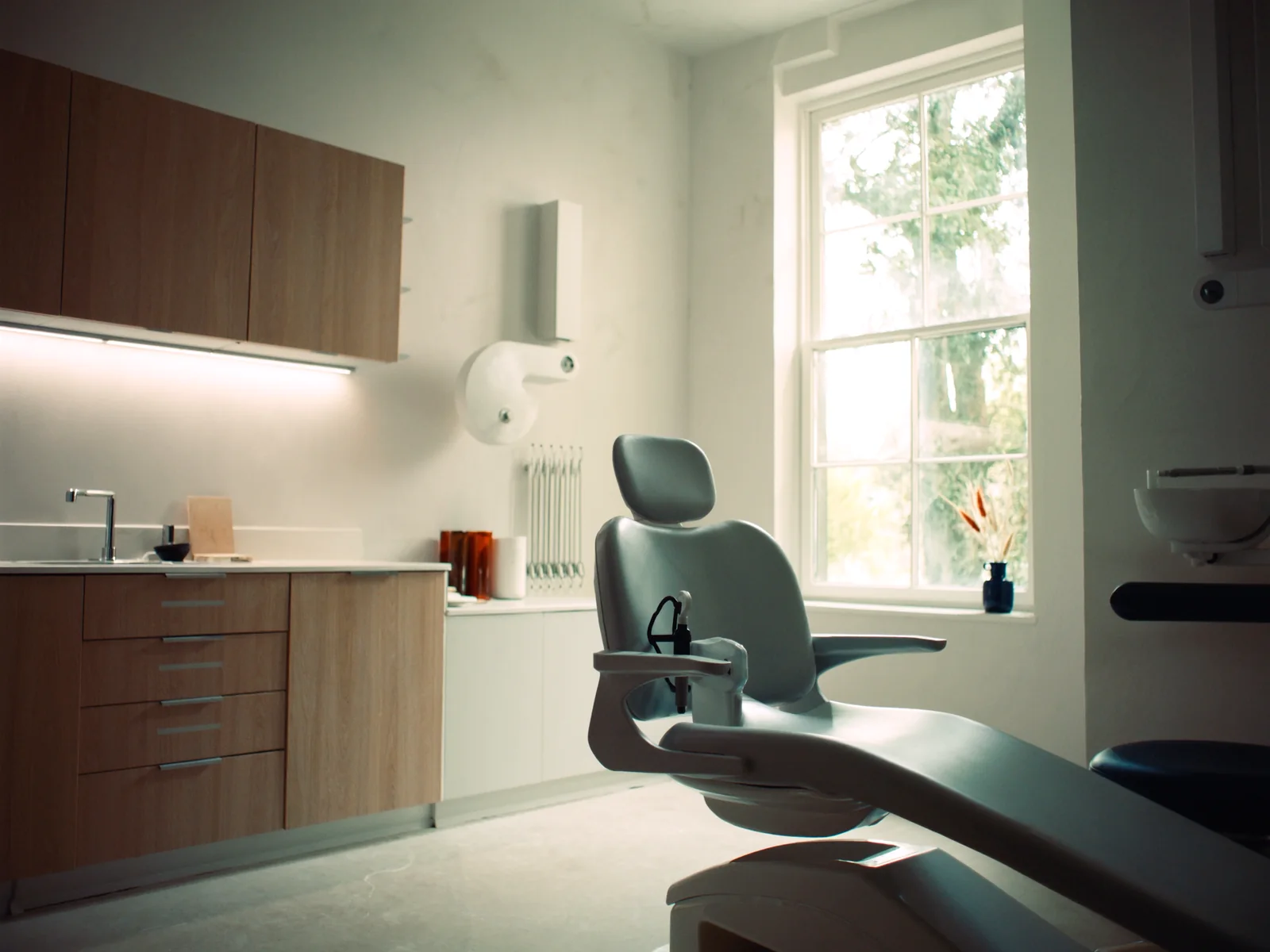 An empty, daylit dental surgery: a treatment chair beside a tall sash window and oak cabinets