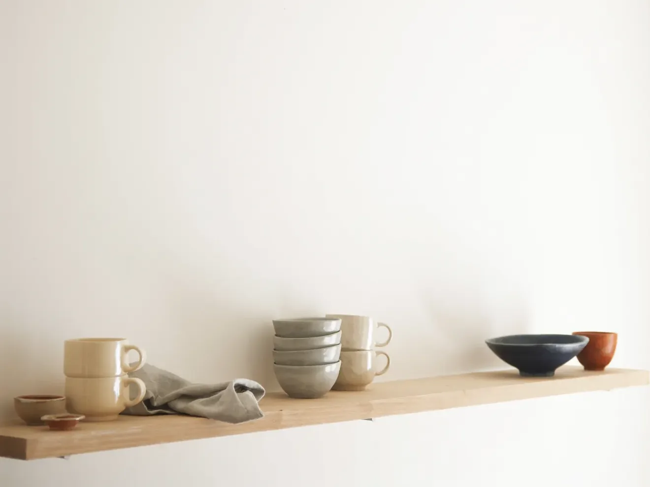 Handmade stoneware cups and bowls on a pale oak shelf against a white wall