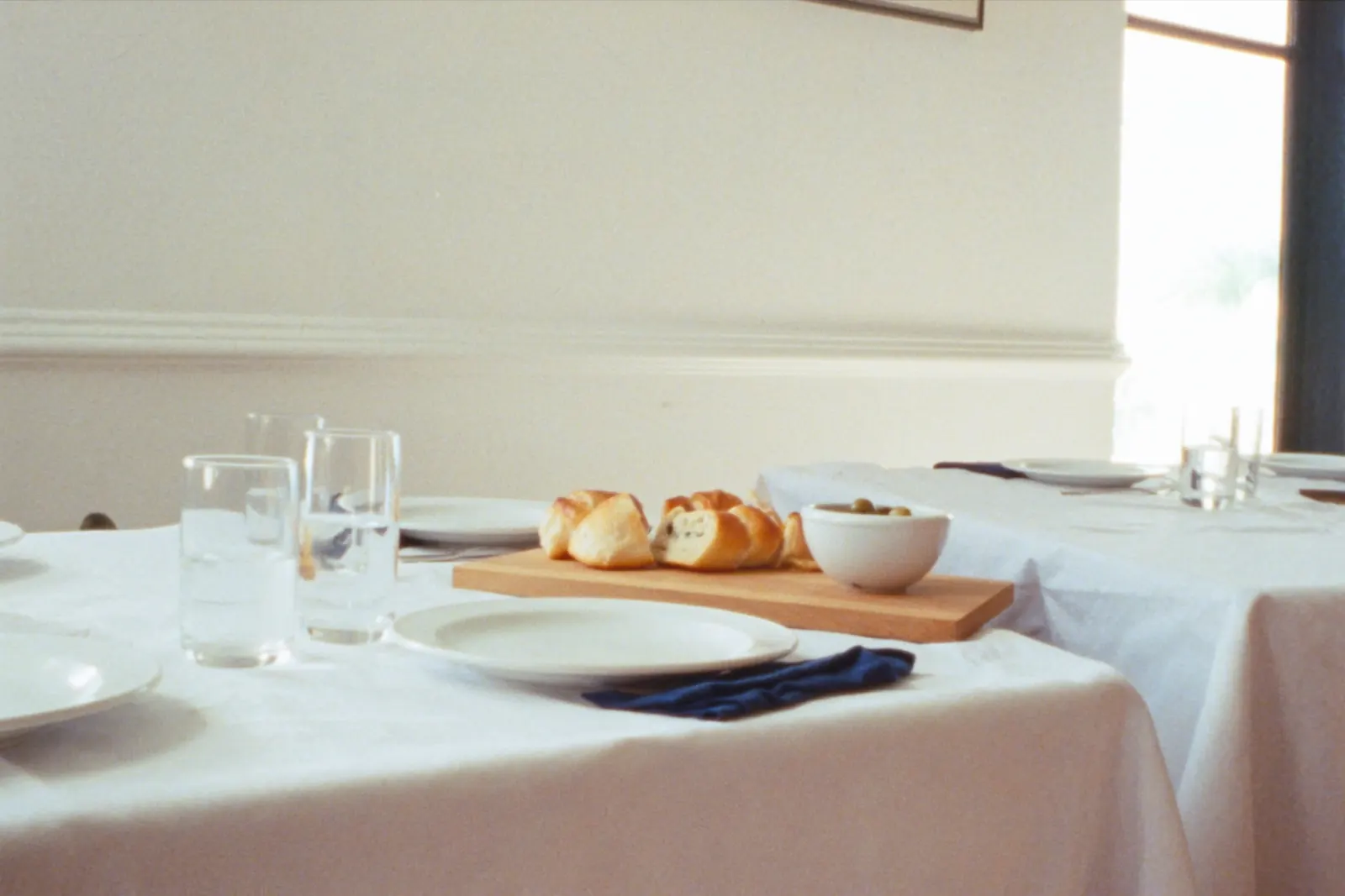 A restaurant table laid for lunch with white linen, plates, glasses and a board of bread
