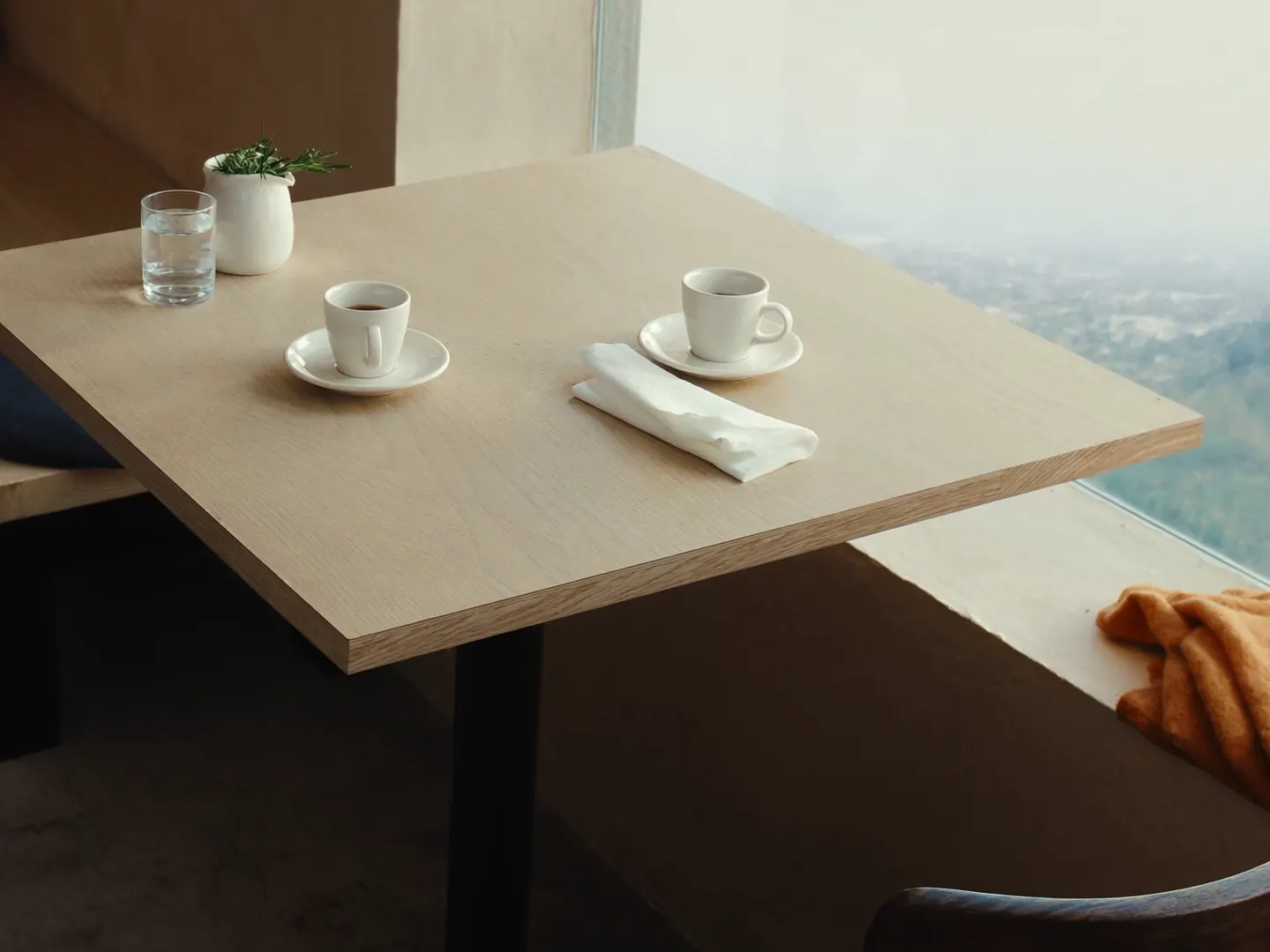 A pale oak café table by a tall window in morning light, set with two espresso cups, a glass of water and a small jug of rosemary