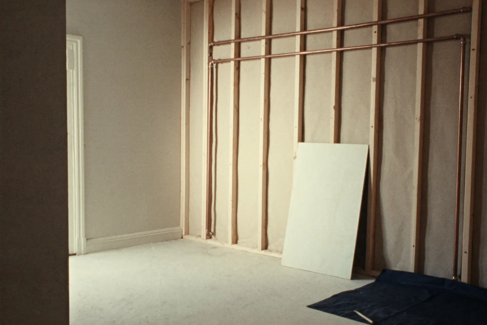 A room mid-renovation: a timber stud wall with runs of copper pipe, a sheet of plasterboard leaning on it and a navy dust sheet on the floor